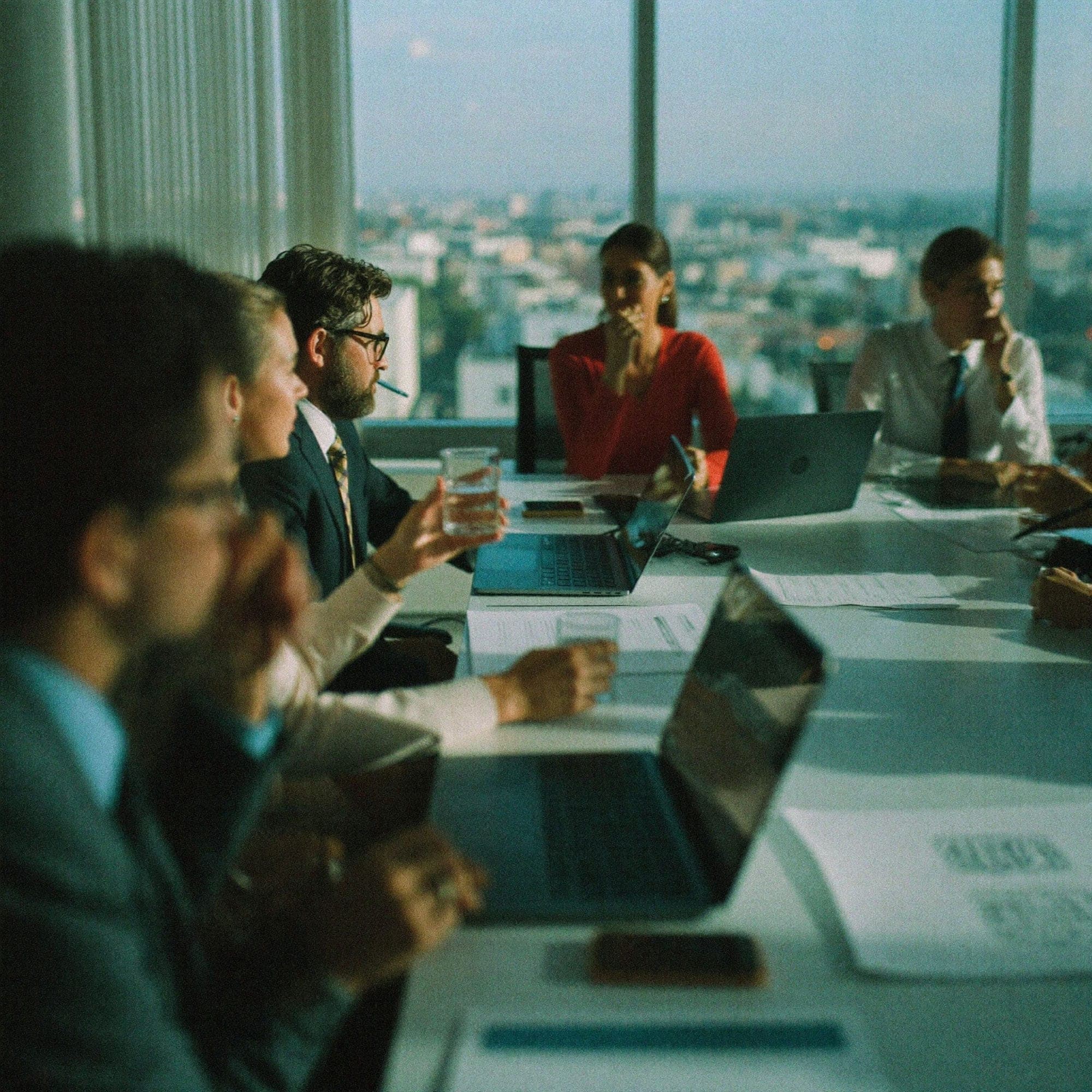 A strategy session in a high-rise boardroom, colleagues around a long table with laptops and documents, the city visible through floor-to-ceiling windows